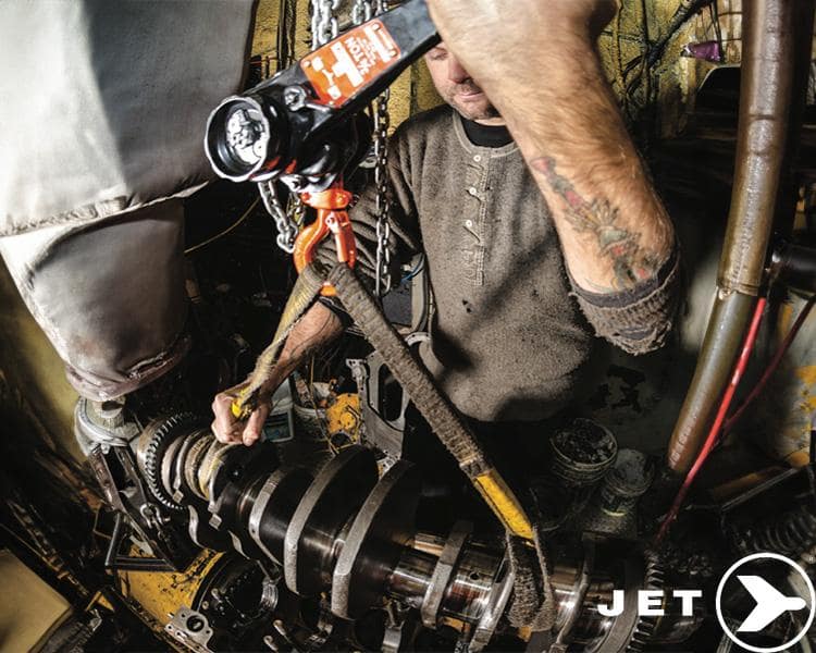 A mechanic with tattooed arms uses a JET Tools hoist to repair industrial machinery in a cluttered workshop.