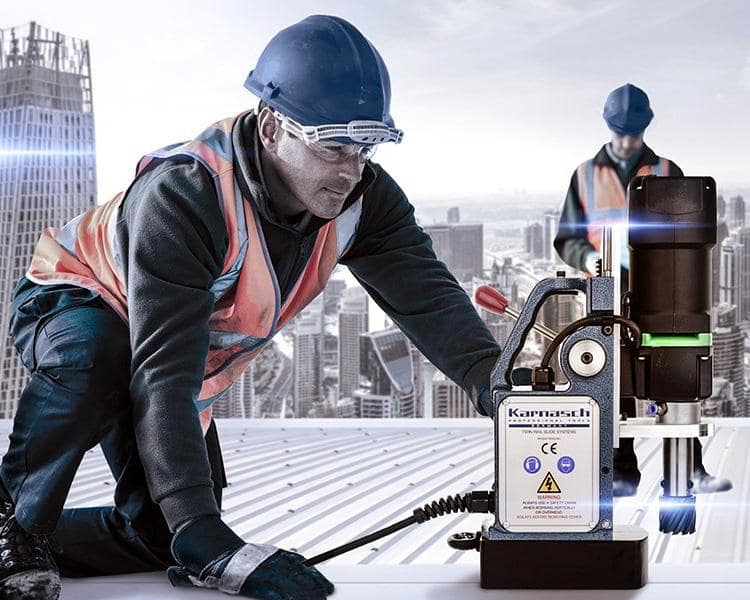 A construction worker wearing a helmet and reflective vest uses survey equipment on a rooftop with a cityscape in the background. Another worker is visible in the distance.