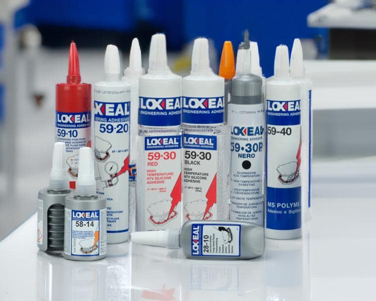 Tubes and bottles of Loctite adhesive products displayed on a table, with focus on items in the foreground against a blurred blue background.