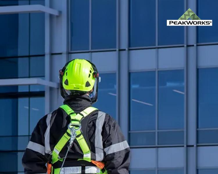 A worker in safety gear, including a helmet and harness, views a large glass building from the outside, with the peakworks logo in the corner.