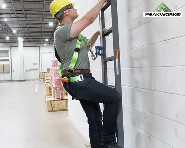 A construction worker in a yellow hard hat and green safety harness is installing equipment on a wall in a warehouse.