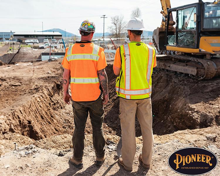 Two construction workers in high visibility vests and hard hats stand near an excavation site with a large bulldozer in the background, seemingly discussing the project.