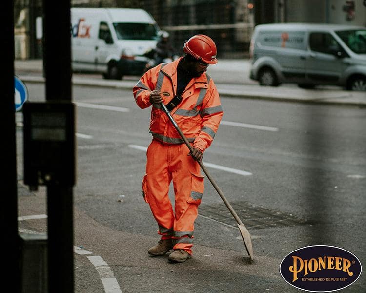 A construction worker in orange reflective gear using a hoe on a city street, with vehicles in the background and a pioneer logo at the bottom right.