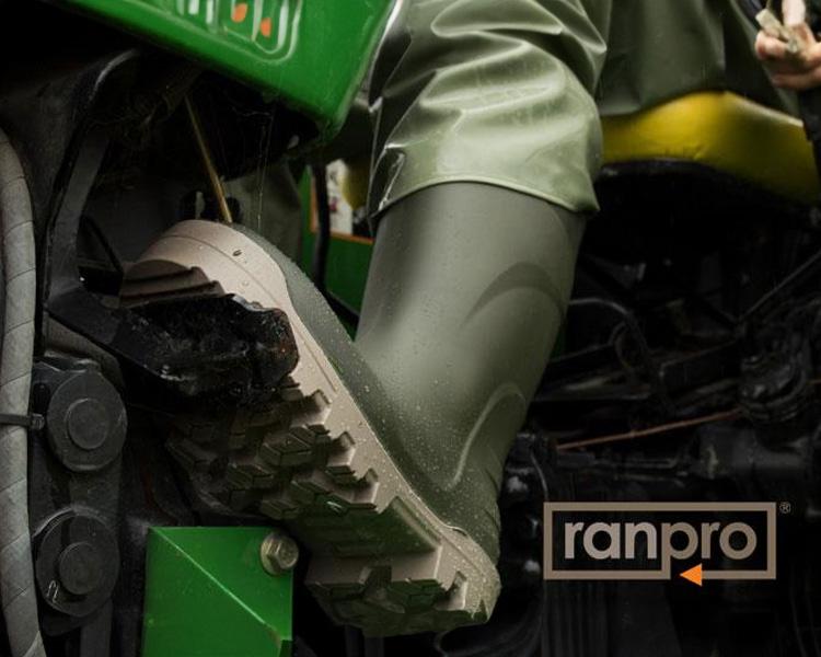 Close-up of a person wearing green rubber boots, pressing a pedal on a green agricultural machine, with visible dirt on the boots and the machine's tire. the ranpro logo is in the corner.