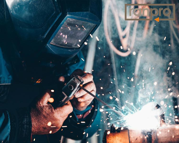 Welder wearing protective gear using a welding torch on metal, with sparks flying around in a dimly lit workshop, with the ranpro logo in the top right corner.