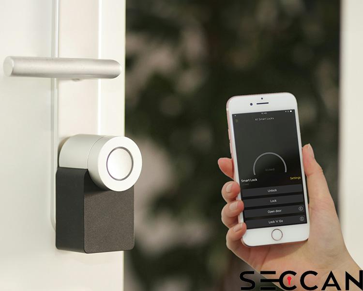 A person's hand holding a smartphone with an app opened to control a Seccan smart lock, which is attached to a door in the background. The app screen shows buttons for locking and unlocking the door.