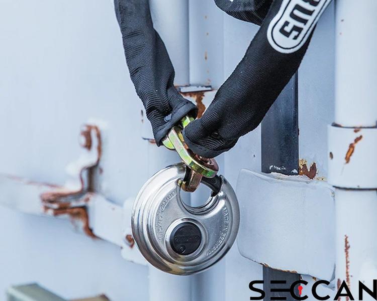 A person wearing black gloves locks a sturdy silver padlock on a rusted metal gate, with the Seccan brand name visible on equipment near the lock.