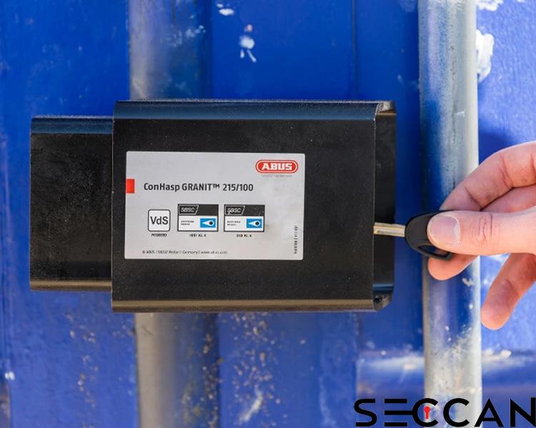 A person's hand inserting a key into an ABUS heavy-duty Granit padlock mounted on a blue metal structure, with Seccan branding visible.